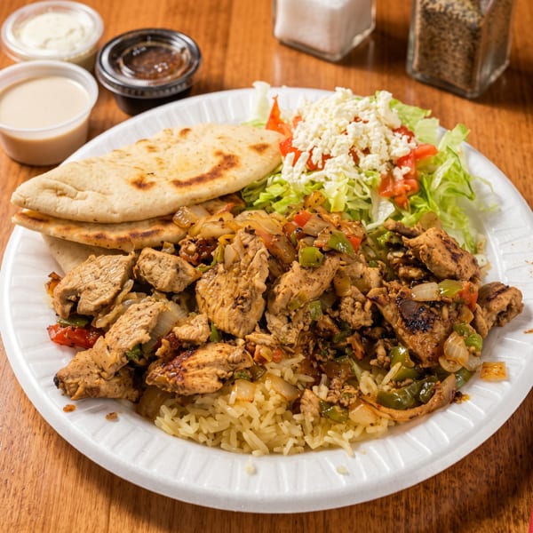 Chicken and rice platter with salad, pita bread, and dipping sauces on a wooden table.