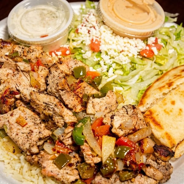 Grilled chicken and vegetables over rice, with a side salad, pita bread, and two dipping sauces.