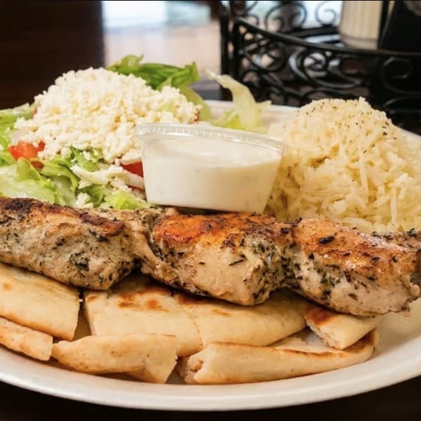 Grilled chicken skewer with pita bread, rice, Greek salad, and a side of tzatziki sauce.